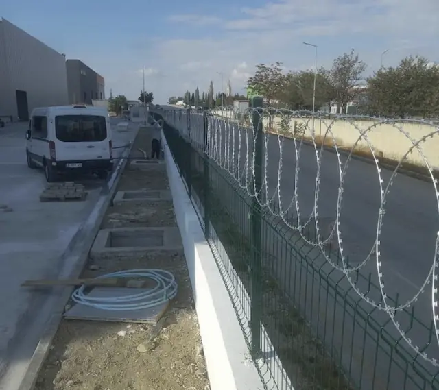 Fabrika önünde beton duvar üstüne yeşil panel çit ve spiral jiletli tel